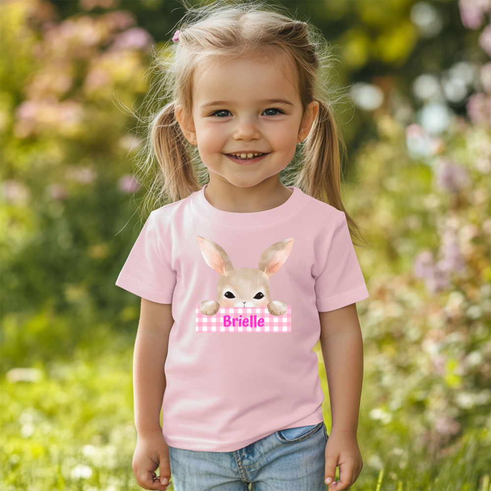 "Personalized kids' Easter t-shirt featuring a cute bunny peeking over a custom name sign. Adorable pastel design, perfect for toddlers and Easter gifts.